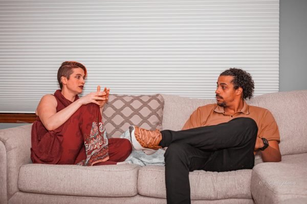 Woman in maroon outfit with red hair sitting on a couch talking to a man in black dress pants, and brown dress shoes and shirt.